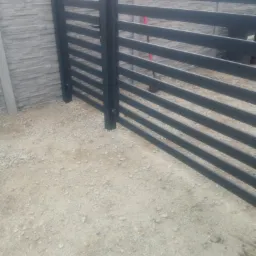 Nowoczesna, pozioma brama wjazdowa z czarnych profili, zamontowana na słupkach z betonu architektonicznego, na podłożu z drobnego żwiru.