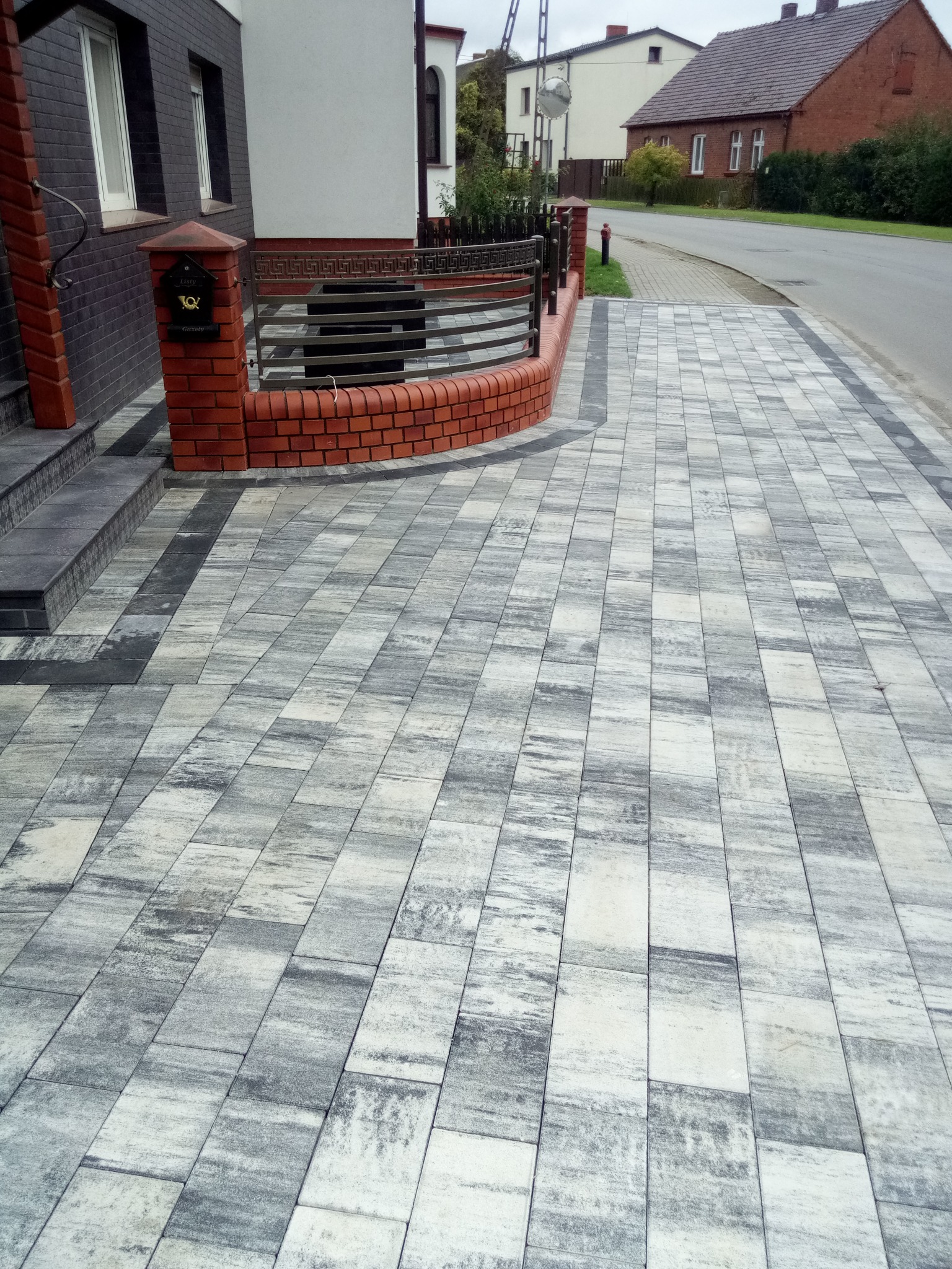 Ułożona kostka brukowa w odcieniach szarości tworząca chodnik przed domem z ceglanym ogrodzeniem i skrzynką na listy. Widok na ulicę i domy w tle.