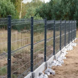 Szary panelowy płot z podmurówką i kamieniami u podstawy, widok z boku, drzewa w tle.