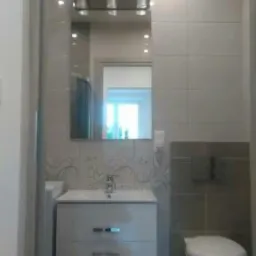 Wykończona łazienka z białą szafką pod umywalką, lustrem z oświetleniem, toaletą w zabudowie i szaro-białymi płytkami z delikatnym wzorem.