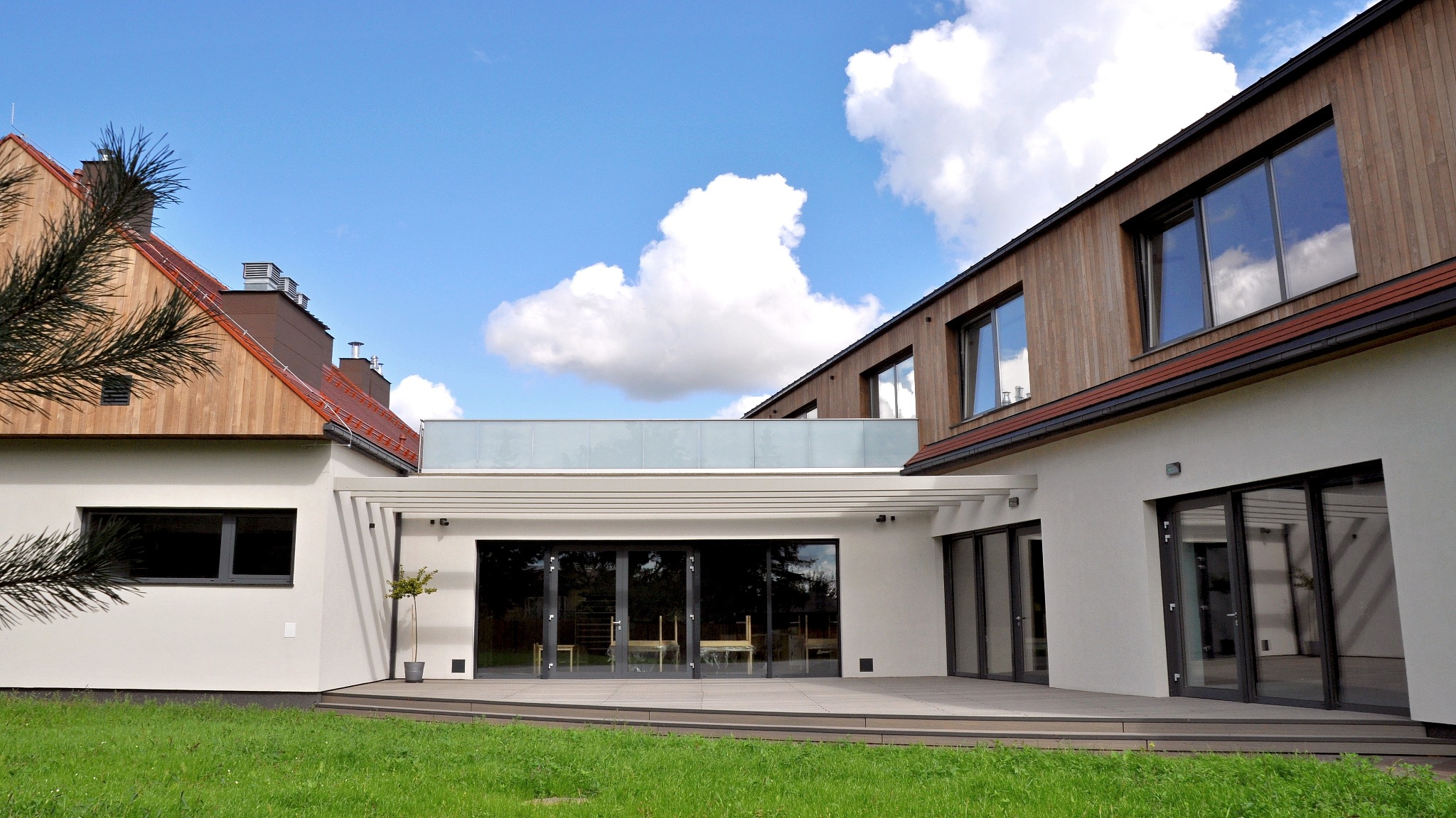 Nowoczesny budynek przedszkola z drewnianą elewacją i dużymi oknami, widok z zewnątrz na tle błękitnego nieba z chmurami.