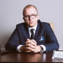 Portret adwokata w garniturze siedzącego za biurkiem z laptopem i wizytówką, splecionymi dłońmi, patrzącego wprost, z powagą w oczach, na neutralnym tle.