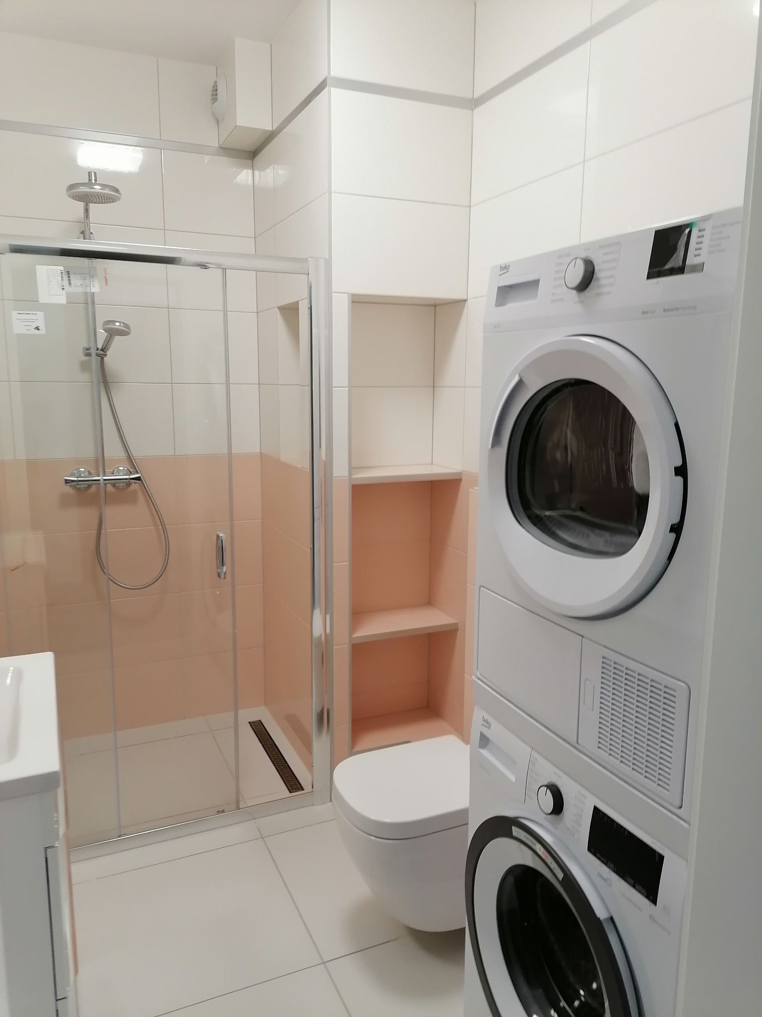 Nowoczesna łazienka z kabiną prysznicową, toaletą i pralko-suszarką Beko ustawioną w pionie, wykończona płytkami w odcieniach bieli i pastelowego różu.