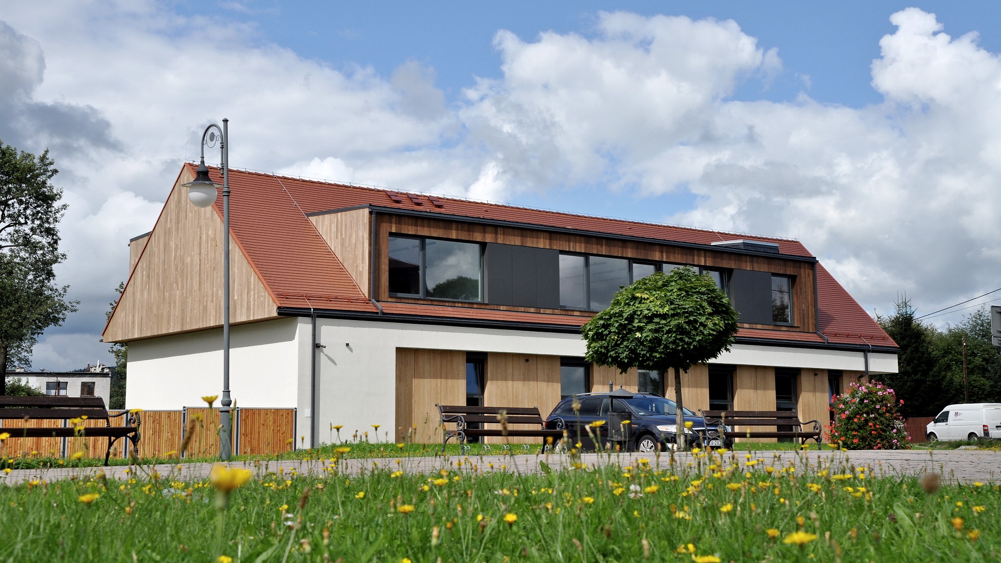 Nowoczesny budynek przedszkola z jasną drewnianą elewacją, czerwoną dachówką i dużymi oknami, otoczony zielonym trawnikiem z żółtymi kwiatami, widok z poziomu gruntu.