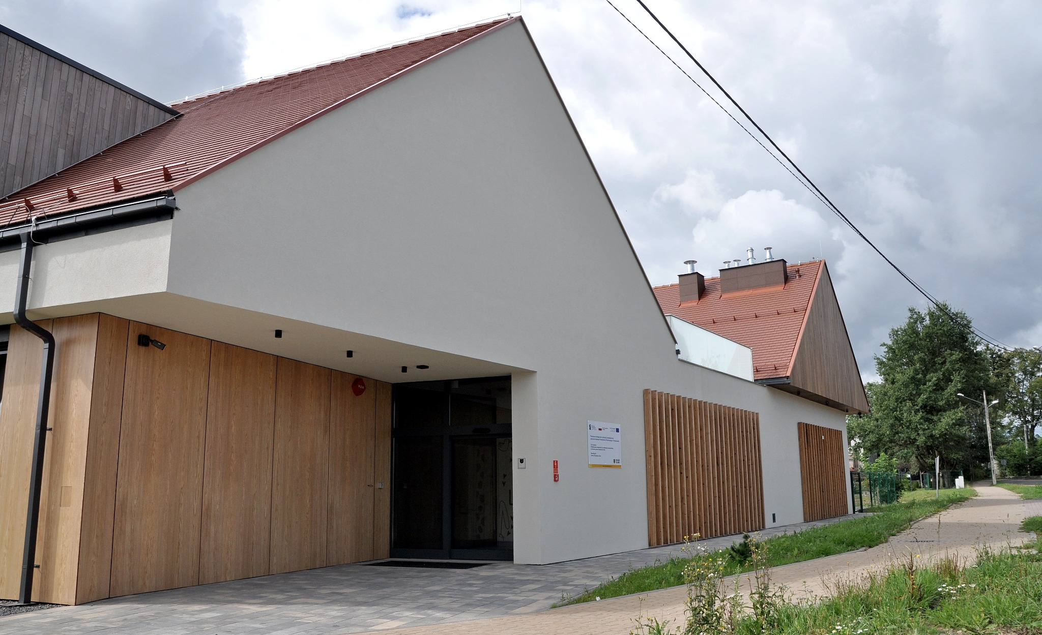Nowoczesny budynek przedszkola publicznego w Krzeszowie, z białymi ścianami, drewnianą elewacją i czerwoną dachówką, zaprojektowany przez architekta Piotra Wyrostka.