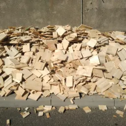 Stos świeżo pociętych, jasnych, drewnianych elementów o kwadratowym i prostokątnym kształcie, leżący na betonowym chodniku przy szarej ścianie, oświetlony jasnym, naturalnym światłem.