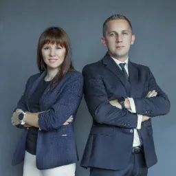 Portret biznesowy: Kobieta w niebieskiej marynarce w kratę i mężczyzna w granatowym garniturze, stoją plecami do siebie z założonymi rękami na tle szarej ściany.