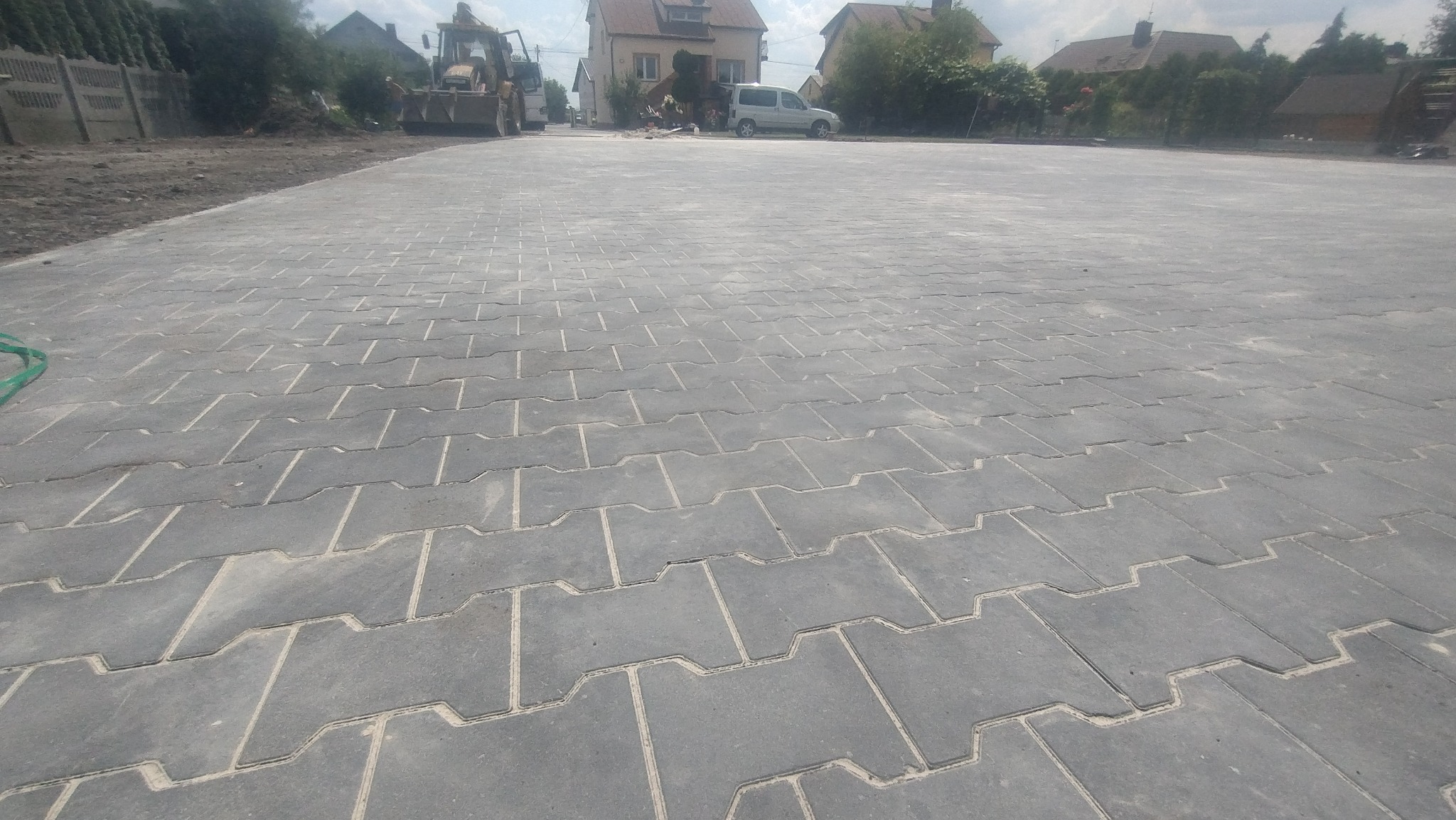 Szeroki plac wyłożony szarą kostką brukową typu 'I', z widocznym traktorem i budynkami w tle, sugerujący świeżo ukończoną realizację.