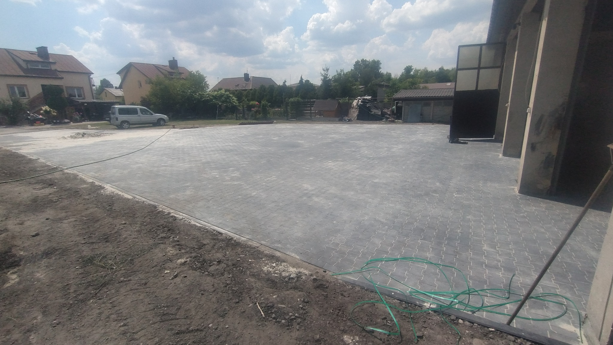 Rozległy, szary plac wyłożony kostką brukową typu holland, z widocznym obrzeżem i częściowo przygotowanym podłożem, w tle zabudowania mieszkalne i samochód.