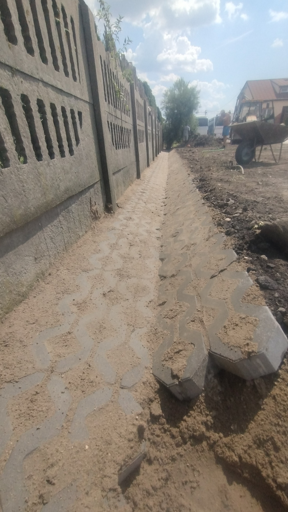 Fragment nowej ścieżki z betonowej kostki o zygzakowatym wzorze, zasypanej piaskiem, przylegającej do betonowego ażurowego ogrodzenia, w tle widoczne prace ziemne i budynki.