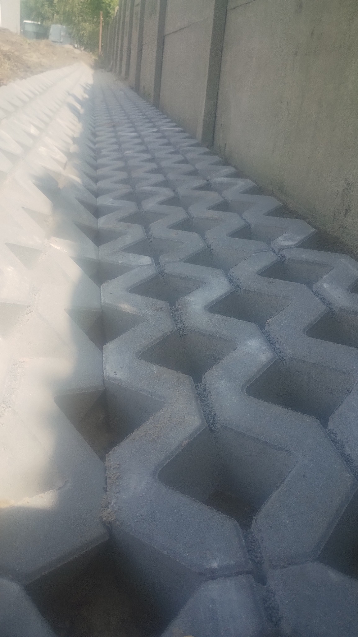 Szara, ażurowa nawierzchnia z betonowych elementów ułożona wzdłuż betonowego muru, wypełniona drobnym kruszywem.