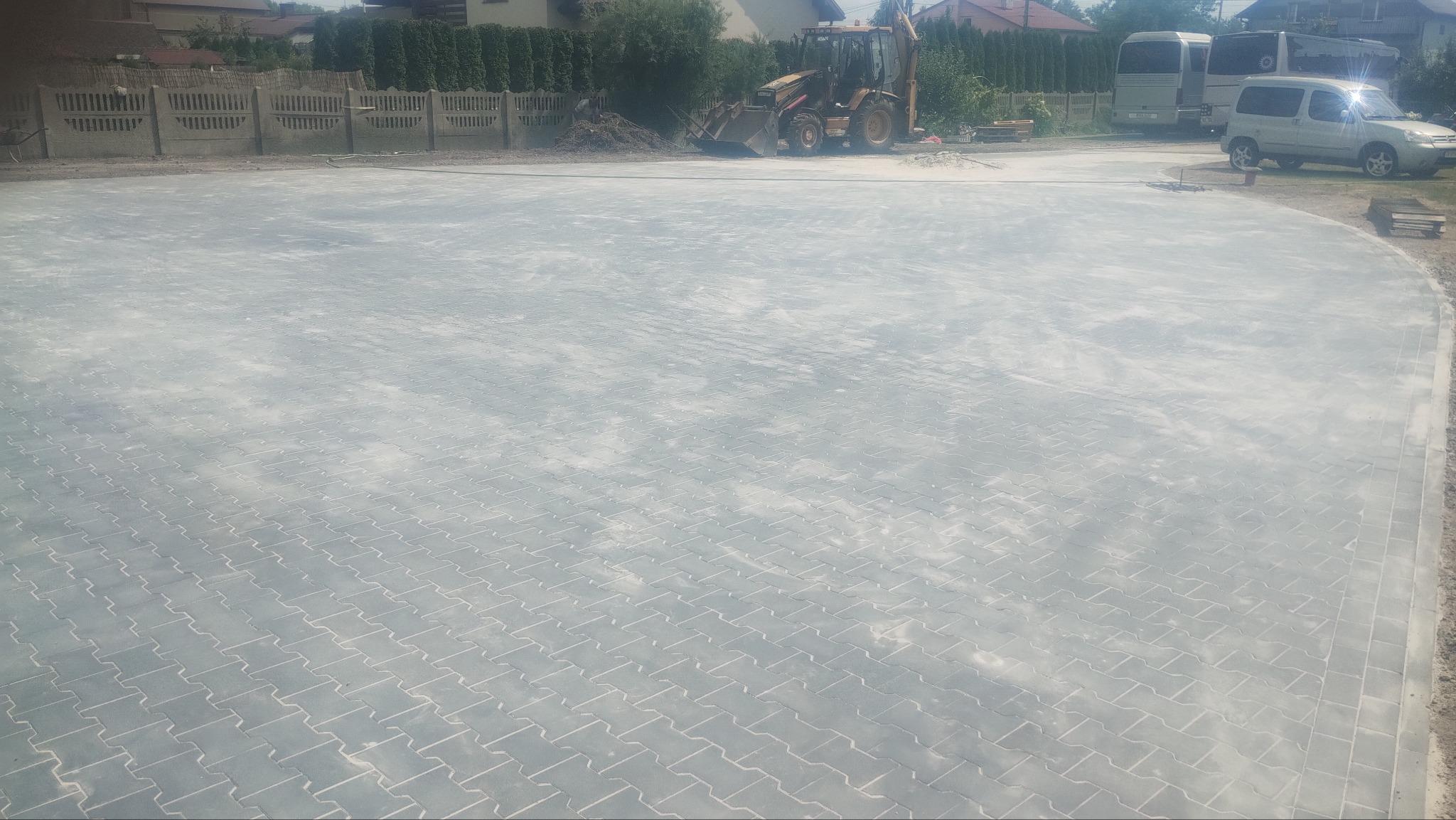 Powierzchnia z ułożonej jasnoszarej kostki betonowej w jodełkę, z koparką w tle i częściowo zaparkowanymi pojazdami, w słoneczny dzień.