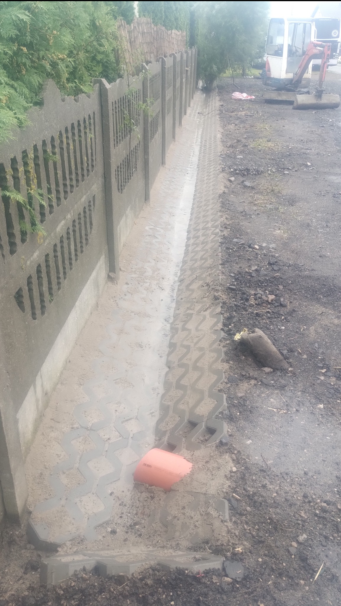 Montaż prefabrykowanego ogrodzenia betonowego, widok na fragment ułożonej nawierzchni z ażurowych płyt betonowych w błocie, pomarańczowa rura kanalizacyjna, w tle minikoparka.