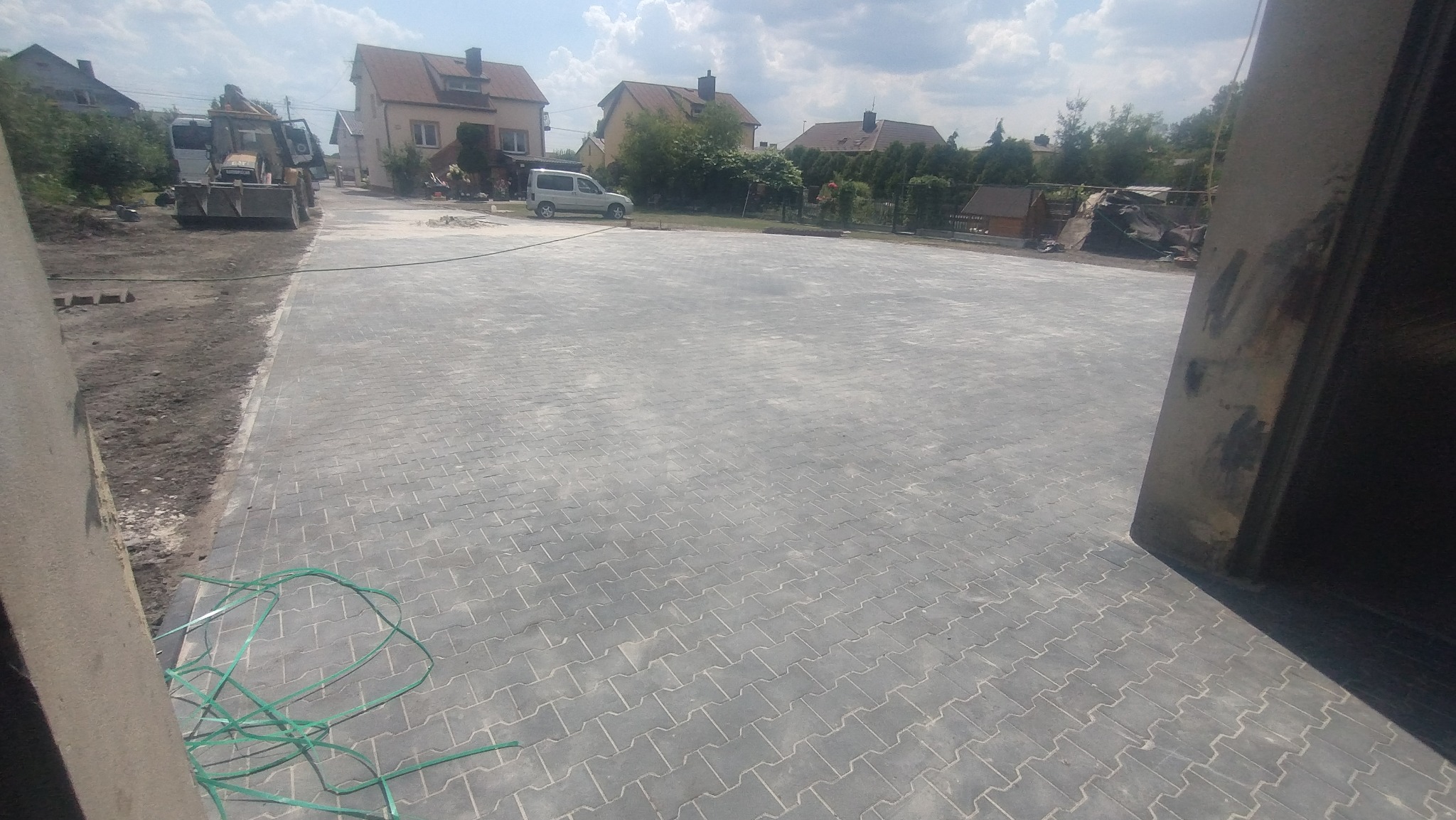 Szeroki plac wyłożony szarą kostką w jodełkę, z widocznym sprzętem budowlanym i zielonym wężem na pierwszym planie, w tle zabudowa jednorodzinna i pochmurne niebo.