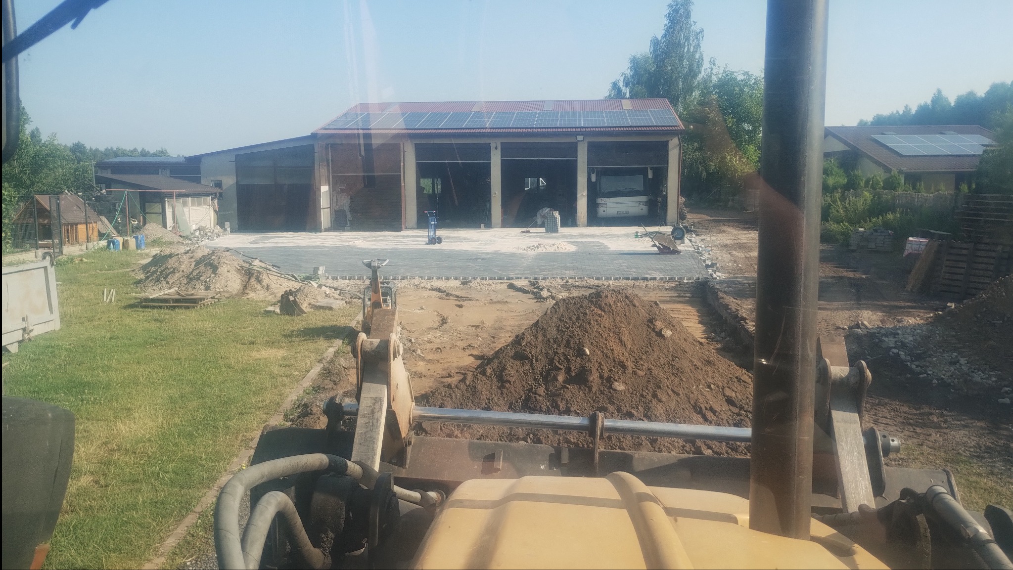 Widok z kabiny maszyny budowlanej na plac budowy z częściowo ułożoną szarą nawierzchnią z kostki, stertą ziemi i budynkiem z panelami słonecznymi w tle.