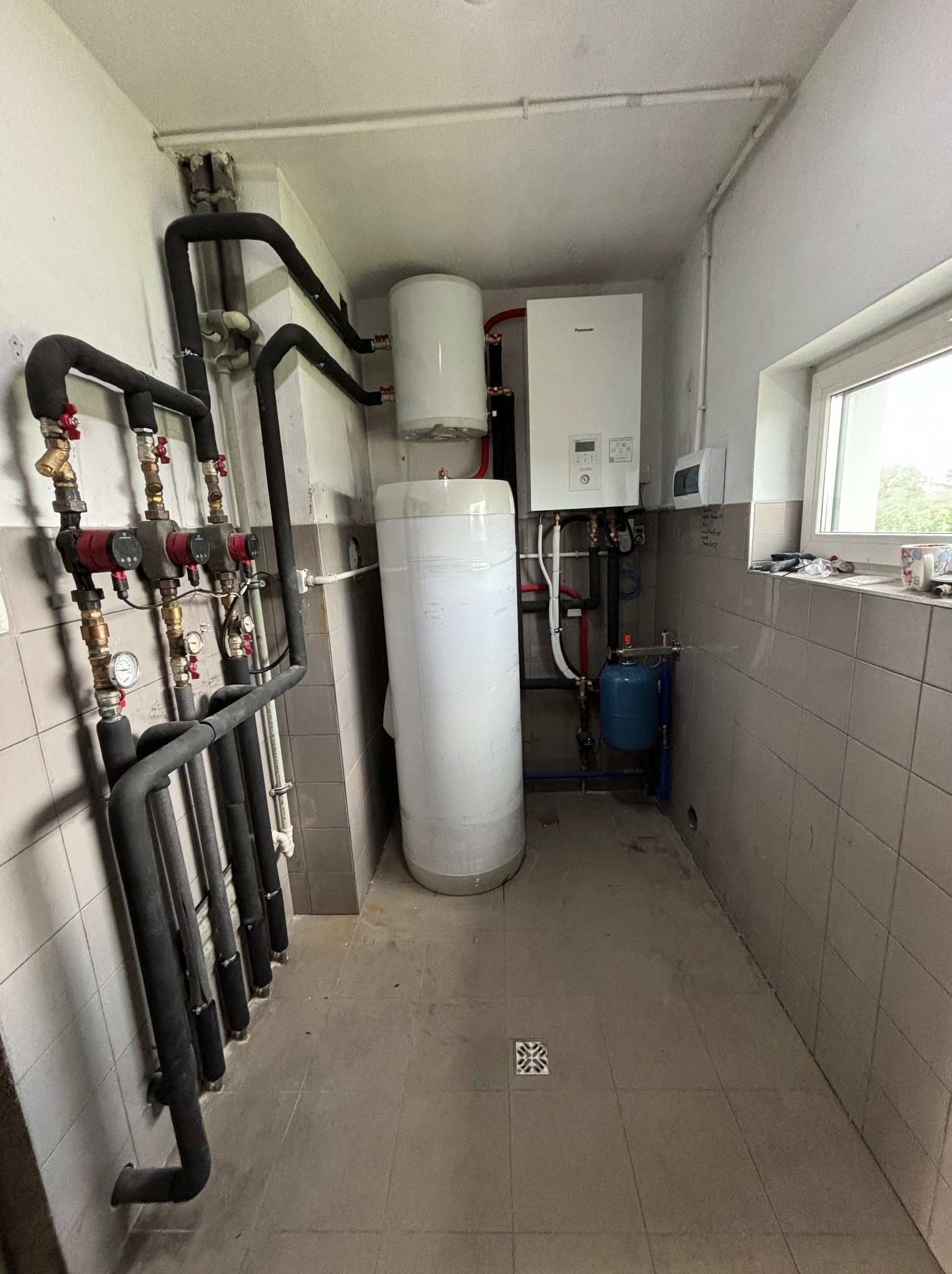 Kotłownia z zamontowaną pompą ciepła Panasonic, zasobnikiem ciepłej wody, grupą pompową z czterema obiegami, instalacją hydrauliczną z rurami w izolacji termicznej i niewielkim zbiornikiem...