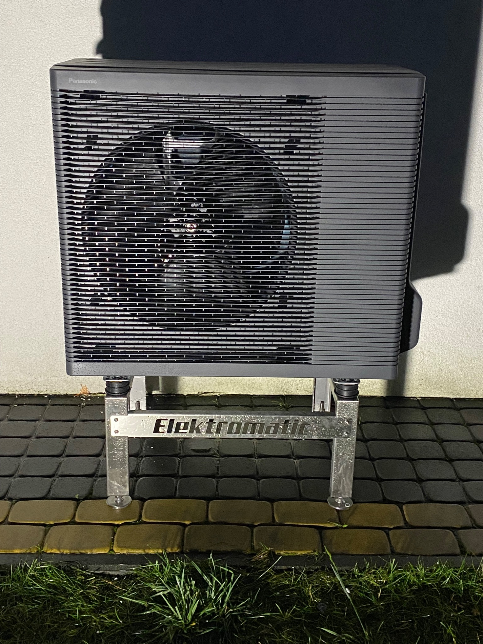 Zewnętrzna jednostka pompy ciepła Panasonic na metalowym stelażu Elektromatic, ustawiona na kostce brukowej, widoczne źdźbła trawy u podstawy.