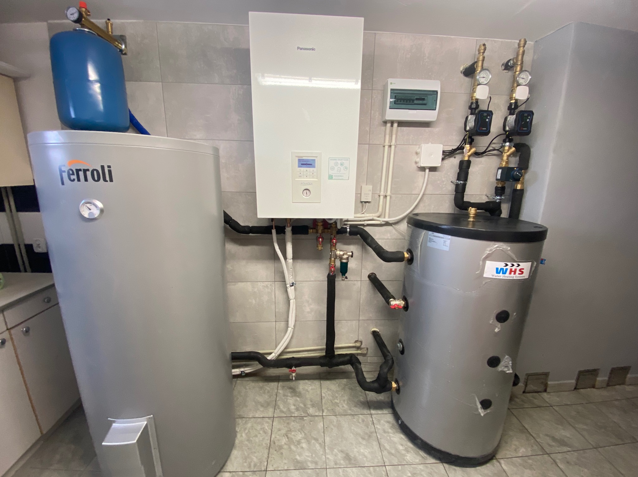Zestaw urządzeń grzewczych w kotłowni: biały piec Panasonic Aquarea Smart Cloud, szary zasobnik Ferroli z niebieskim naczyniem przeponowym oraz szary zasobnik WHS, połączone rurami w izolacji,...