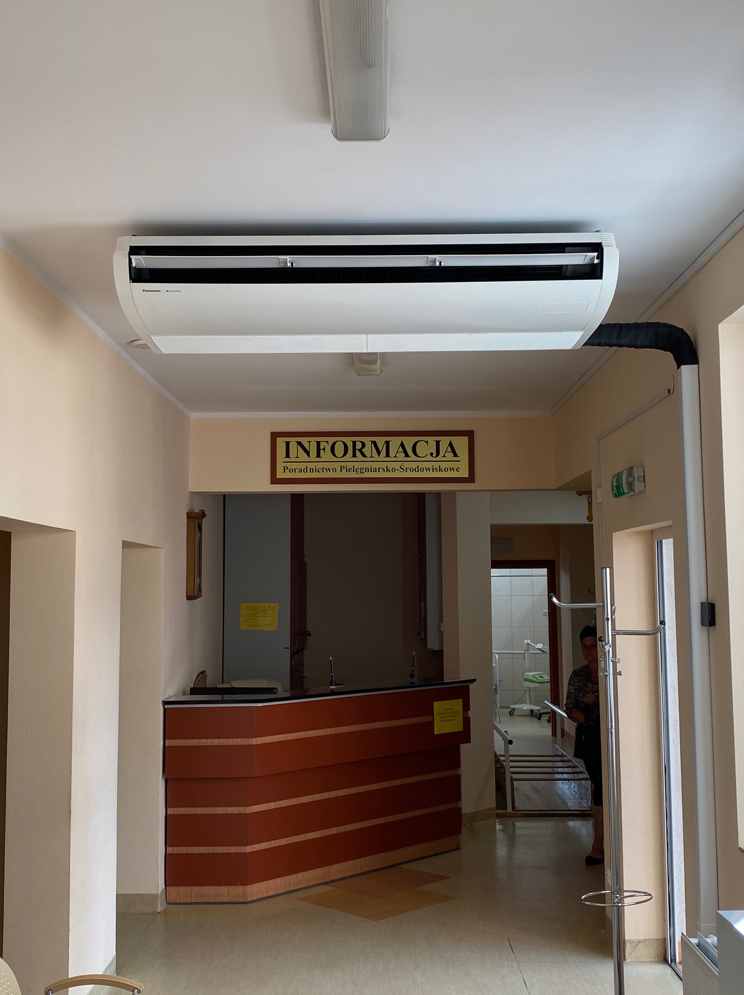 Wnętrze placówki medycznej z zamontowanym klimatyzatorem kasetonowym pod sufitem, widoczny czarny przewód odprowadzający skropliny, na ścianie tabliczka informacyjna.