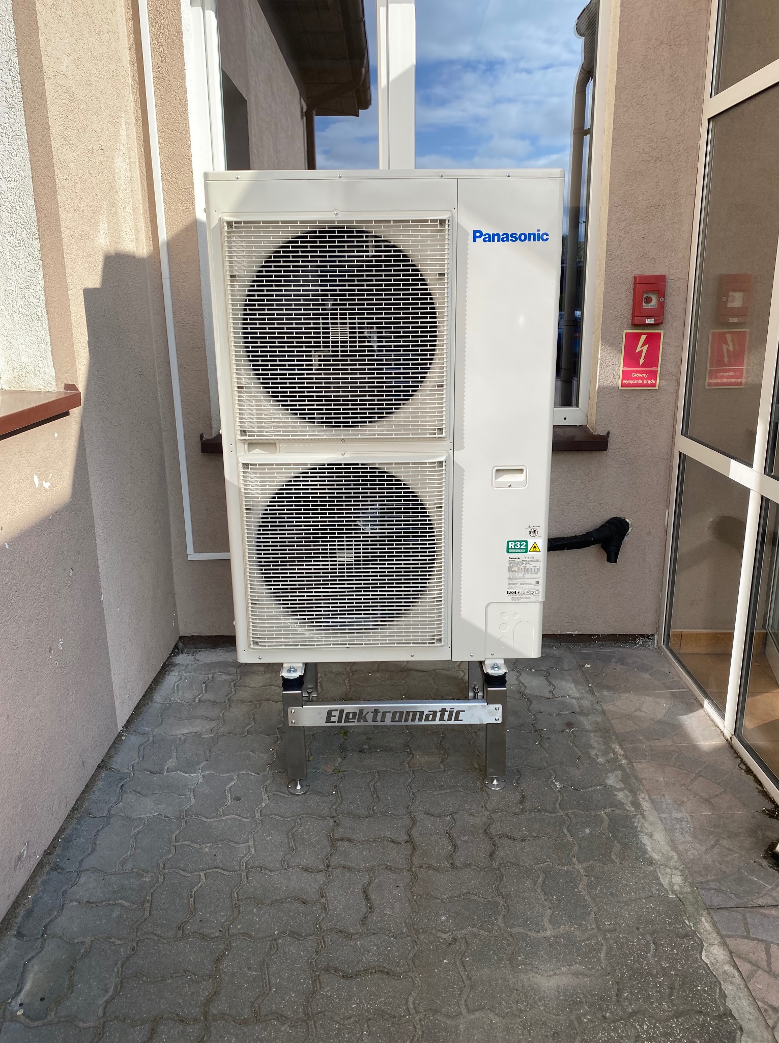 Zewnętrzna jednostka pompy ciepła Panasonic zamontowana na metalowym stelażu Elektromatic przed budynkiem z widocznym oknem i tabliczką ostrzegawczą.