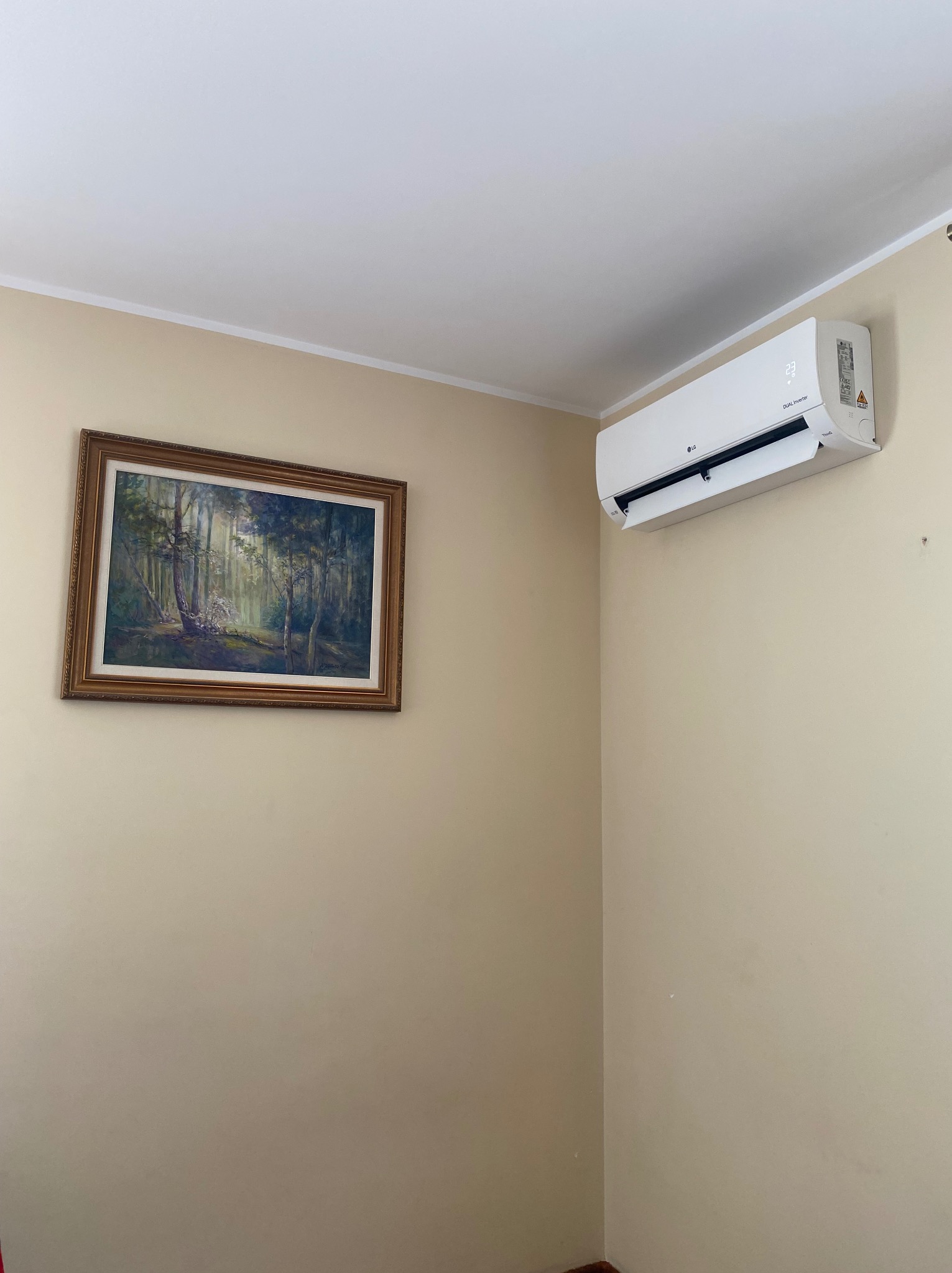 Wnętrze pokoju z zamontowaną na ścianie białą jednostką klimatyzacyjną LG Dual Inverter, ustawioną nad obrazem w złotej ramie przedstawiającym leśny krajobraz.