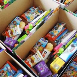 Kartonowe przegródki wypełnione różnorodnymi słodyczami i przekąskami, w tym batoniki, ciastka i napoje w kartonikach, tworzące gotowe zestawy prezentowe.