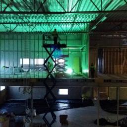 Firma Budowlana LeveL - Wnętrze hali w trakcie budowy z widocznymi elementami konstrukcji stalowej, pracownikami na podnośnikach i charakterystycznym zielonym oświetleniem.