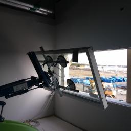 Firma Budowlana LeveL - Montaż okna za pomocą specjalistycznego ramienia ssącego w surowym wnętrzu, widok na plac budowy z maszynami i kontenerami.