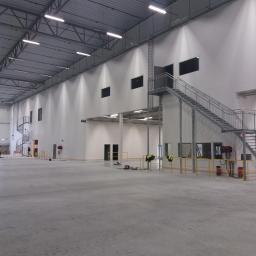 Firma Budowlana LeveL - Wnętrze hali produkcyjnej w trakcie budowy, widoczna konstrukcja stalowa dachu, ściany z oknami, schody stalowe oraz pracownicy na podnośniku nożycowym i na podłodze.