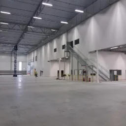 Wnętrze nowo budowanej hali z widoczną konstrukcją stalową dachu, betonową posadzką, białymi ścianami, schodami metalowymi, oknami i pracownikami w oddali.