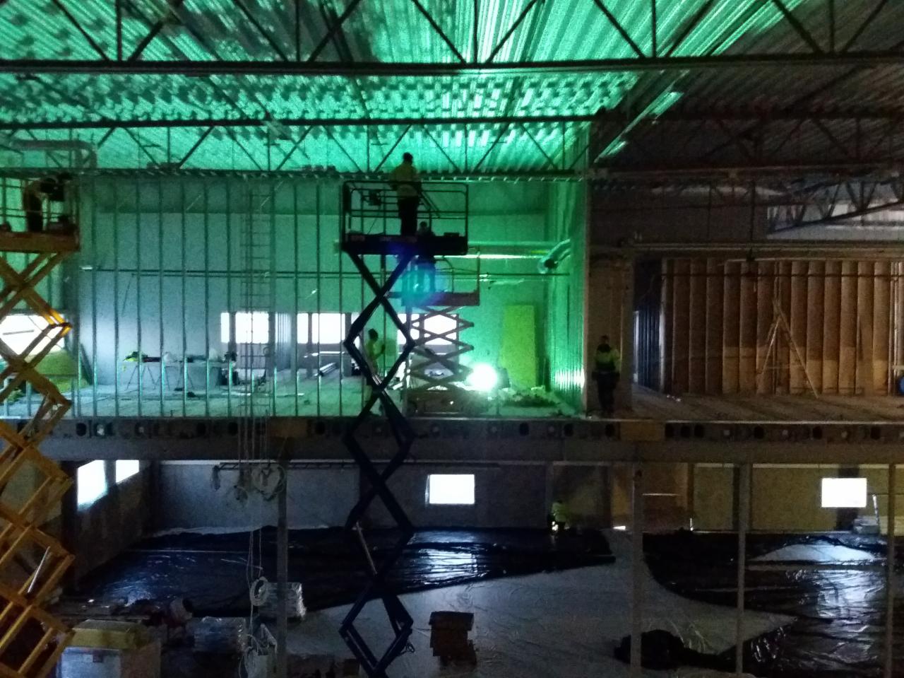 Wnętrze hali w trakcie budowy z widocznymi elementami konstrukcji stalowej, pracownikami na podnośnikach i charakterystycznym zielonym oświetleniem.