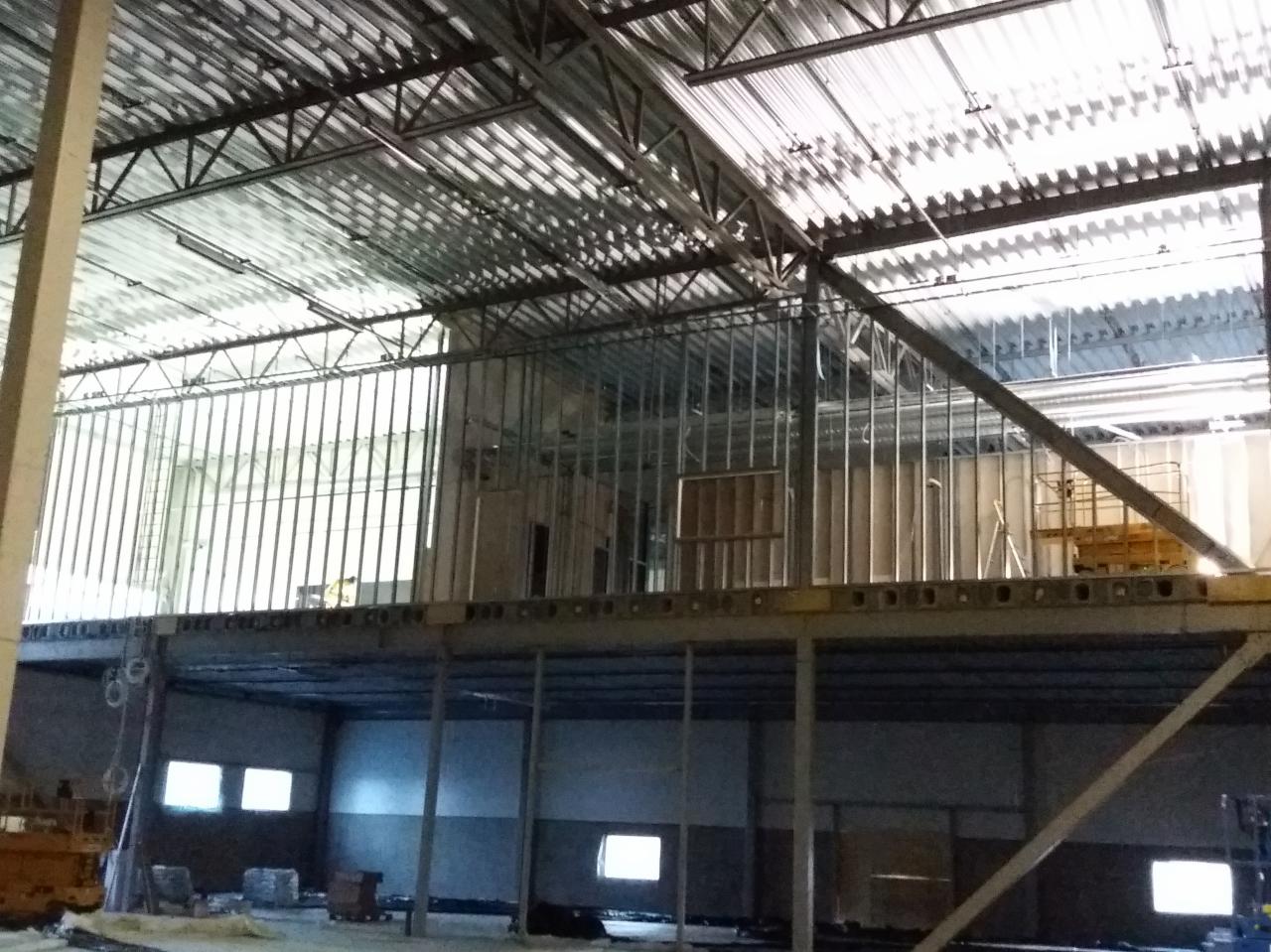 Wnętrze hali w trakcie budowy z widoczną konstrukcją stalową dachu, metalowymi słupami i ścianami działowymi na antresoli, oświetlone światłem dziennym wpadającym przez okna.