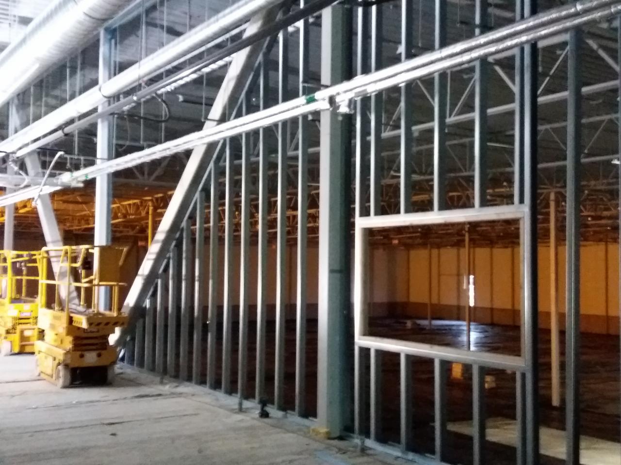 Wnętrze hali w trakcie budowy, widoczne stalowe konstrukcje szkieletowe, dźwig podnośnikowy oraz instalacje pod sufitem.