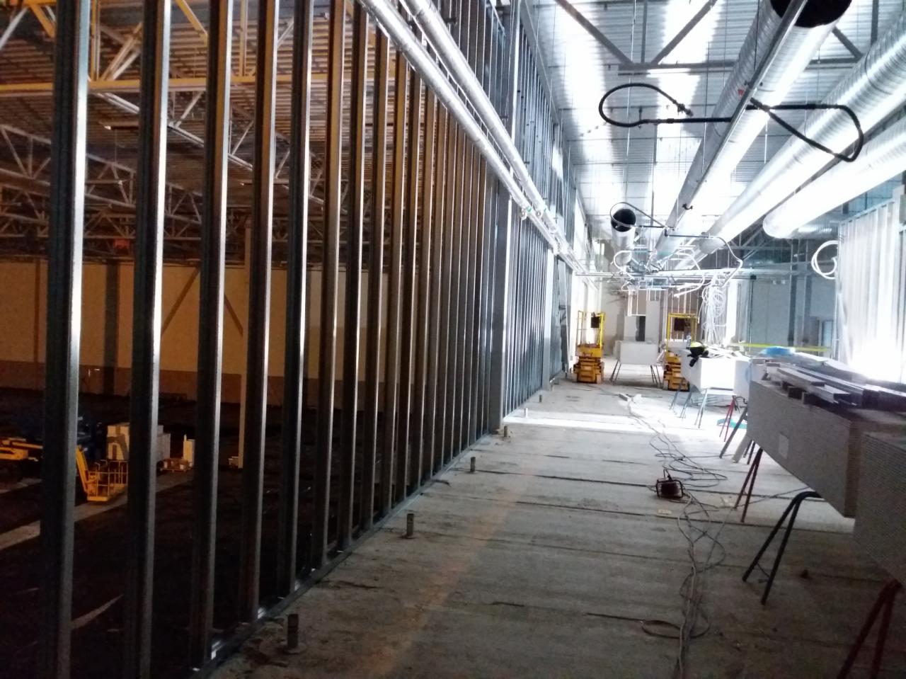 Konstrukcja szkieletowa ścian z profili stalowych, widoczne pionowe słupki i poziome prowadnice, w tle instalacje pod sufitem i podnośniki nożycowe w hali budowlanej.