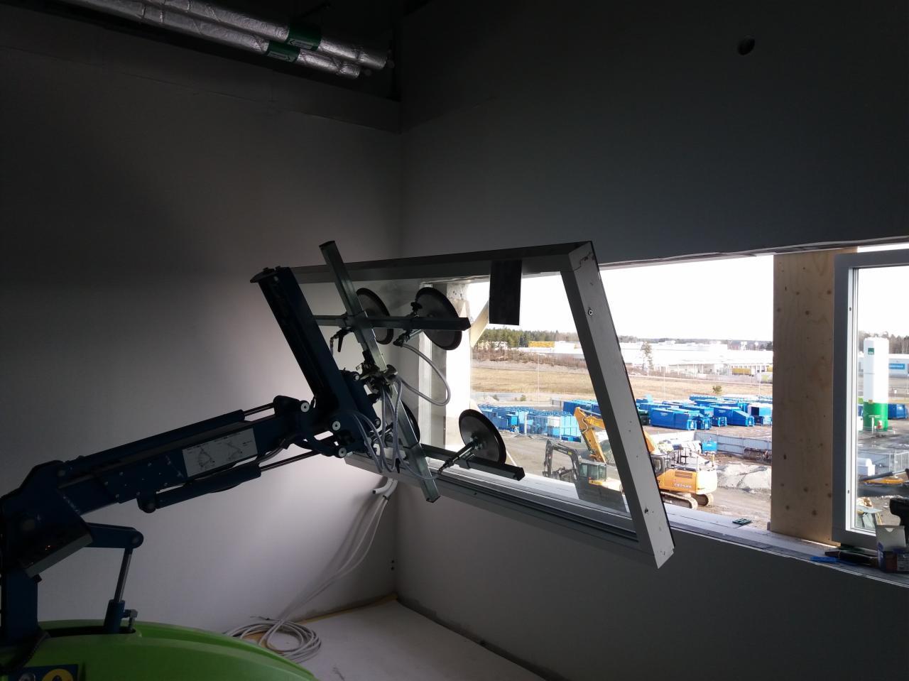 Montaż okna za pomocą specjalistycznego ramienia ssącego w surowym wnętrzu, widok na plac budowy z maszynami i kontenerami.