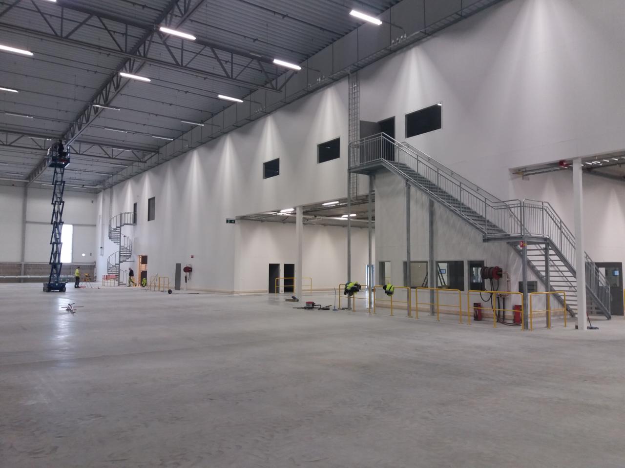 Wnętrze hali produkcyjnej w trakcie budowy, widoczna konstrukcja stalowa dachu, ściany z oknami, schody stalowe oraz pracownicy na podnośniku nożycowym i na podłodze.