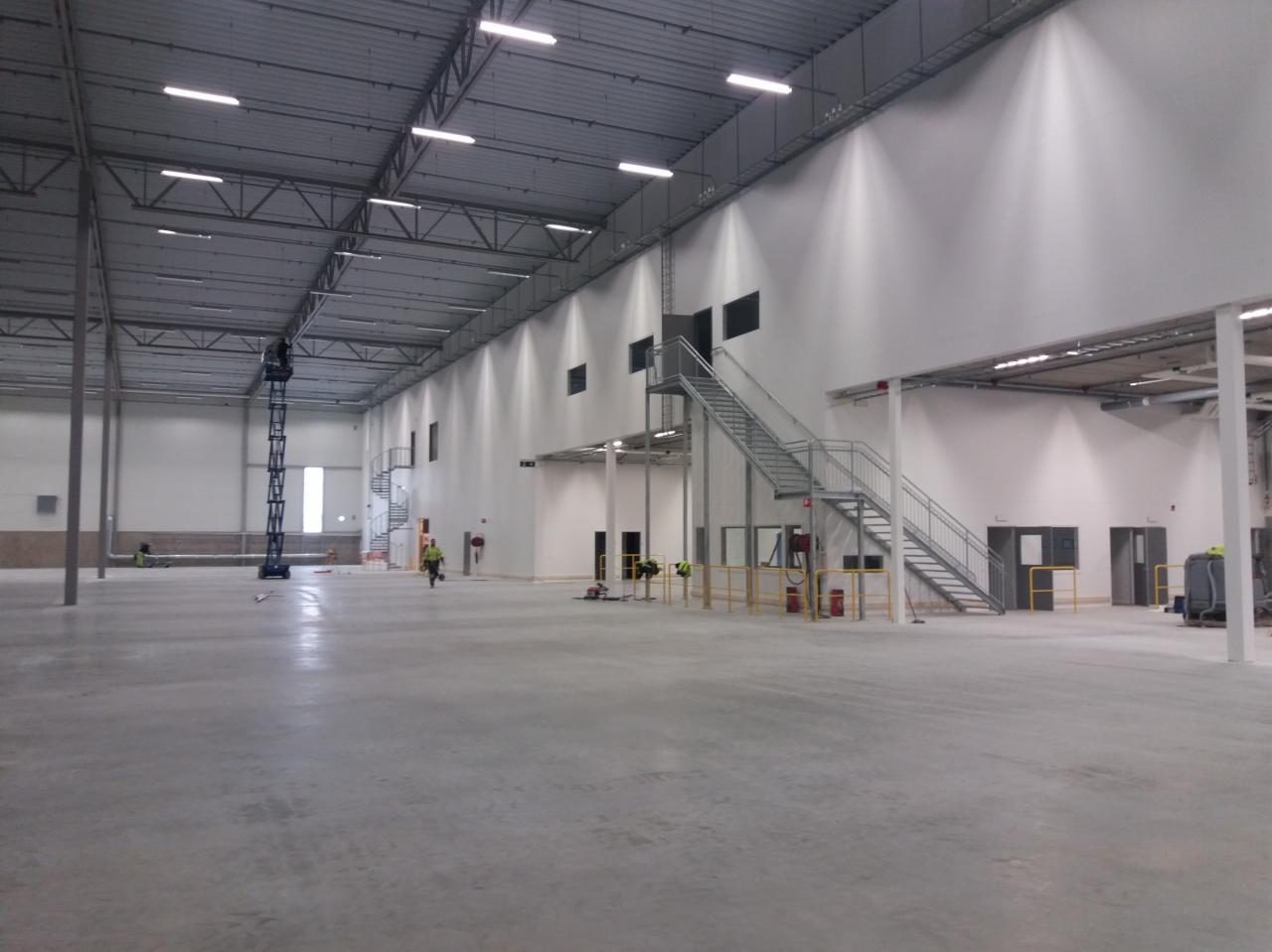 Wnętrze nowo budowanej hali z widoczną konstrukcją stalową dachu, betonową posadzką, białymi ścianami, schodami metalowymi, oknami i pracownikami w oddali.