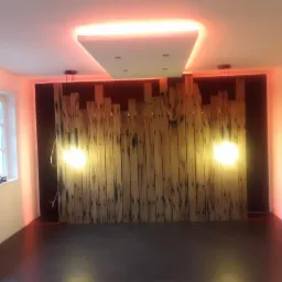 Pokój z dekoracyjną ścianą z surowego drewna podświetloną dwoma wiszącymi lampami, dodatkowo oświetlenie LED na suficie tworzy ciepłą, pomarańczową poświatę na ścianach.