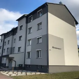 Odnowiony blok mieszkalny z elewacją w odcieniach szarości i bieli, z widocznym adresem 'Klasztorna 6' oraz zainstalowanym monitoringiem.