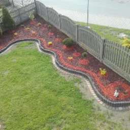 Widok z góry na świeżo zagospodarowany ogród z rabatami wysypanymi korą, obrzeżami z betonowych palisad i elementami dekoracyjnymi w postaci figurek.