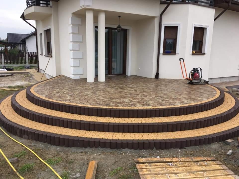 Wykończony taras z dwukolorowej kostki brukowej ułożonej w półokrągłe schody przed domem, z zagęszczarką w tle.