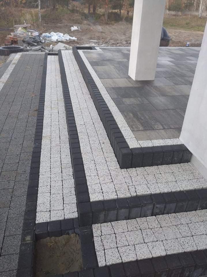 Układanie jasnoszarej kostki betonowej z ciemnoszarą obwódką na schodach i chodniku, widoczny fragment niedokończonej nawierzchni z piaskiem.
