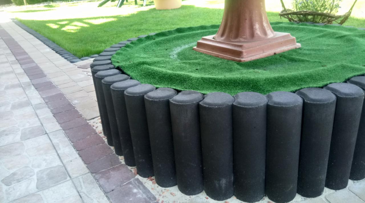 Obrzeże rabaty z czarnych, betonowych palisad, otaczające okrągłą powierzchnię z zieloną sztuczną trawą, na której stoi brązowa podstawa pod donicę lub rzeźbę; obok ułożona kostka brukowa w dwóch...