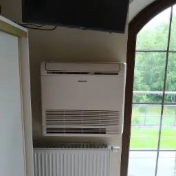 Biały klimatyzator Samsung zamontowany nad grzejnikiem w pokoju z oknem łukowym, widok na zieleń za oknem.