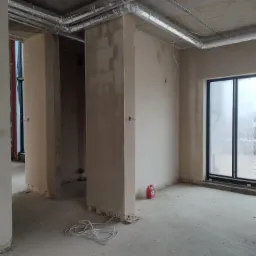 Wnętrze w stanie deweloperskim z nałożonymi tynkami, widoczne przewody elektryczne na podłodze, wentylacja pod sufitem i czerwony kanister.