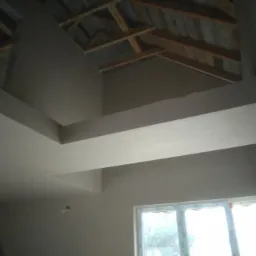 Wnętrze domu w trakcie wykańczania, widoczne gładkie ściany, sufit z drewnianą konstrukcją i fragment okna z widokiem na zewnątrz.