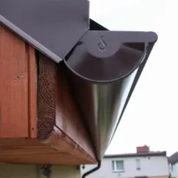 Zamontowana, brązowa rynna z widocznym logo producenta na tle drewnianej podbitki dachowej i fragmentu zabudowań.