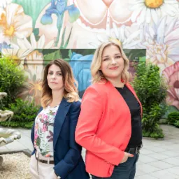 Dwie kobiety w marynarkach stojące tyłem do siebie na tle ściany z muralem kwiatowym i fontanną po lewej stronie.