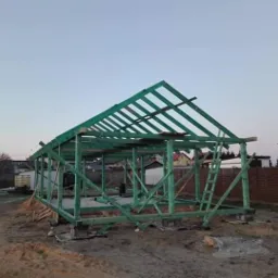 Konstrukcja drewniana dachu w trakcie budowy, zielona impregnacja, widoczna drabina oparta o szkielet, w tle zabudowa jednorodzinna.