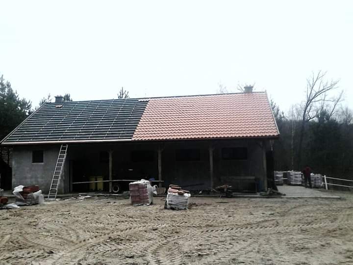 Dom w trakcie krycia dachu: połowa dachu z łatami, druga połowa pokryta czerwoną dachówką, drabina oparta o ścianę, palety z dachówką na ziemi.